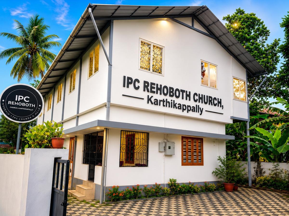 The IPC Rehoboth Church building in Karthikappally — a white two-storey building with the church's name on the wall, lit warmly at dusk beside a coconut palm